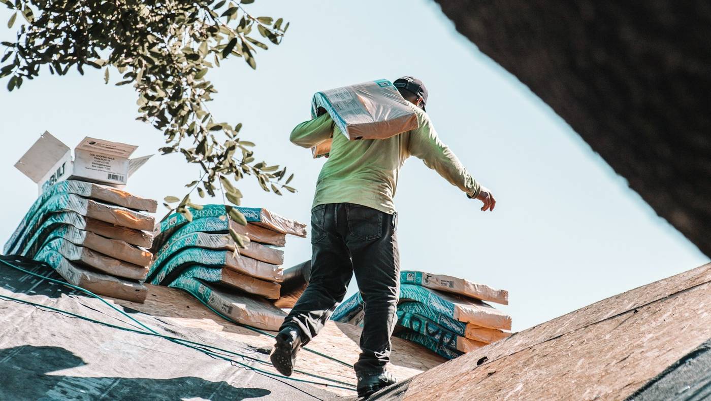 Roofer carrying a bundle of shingles across a roof deck with materials staged
